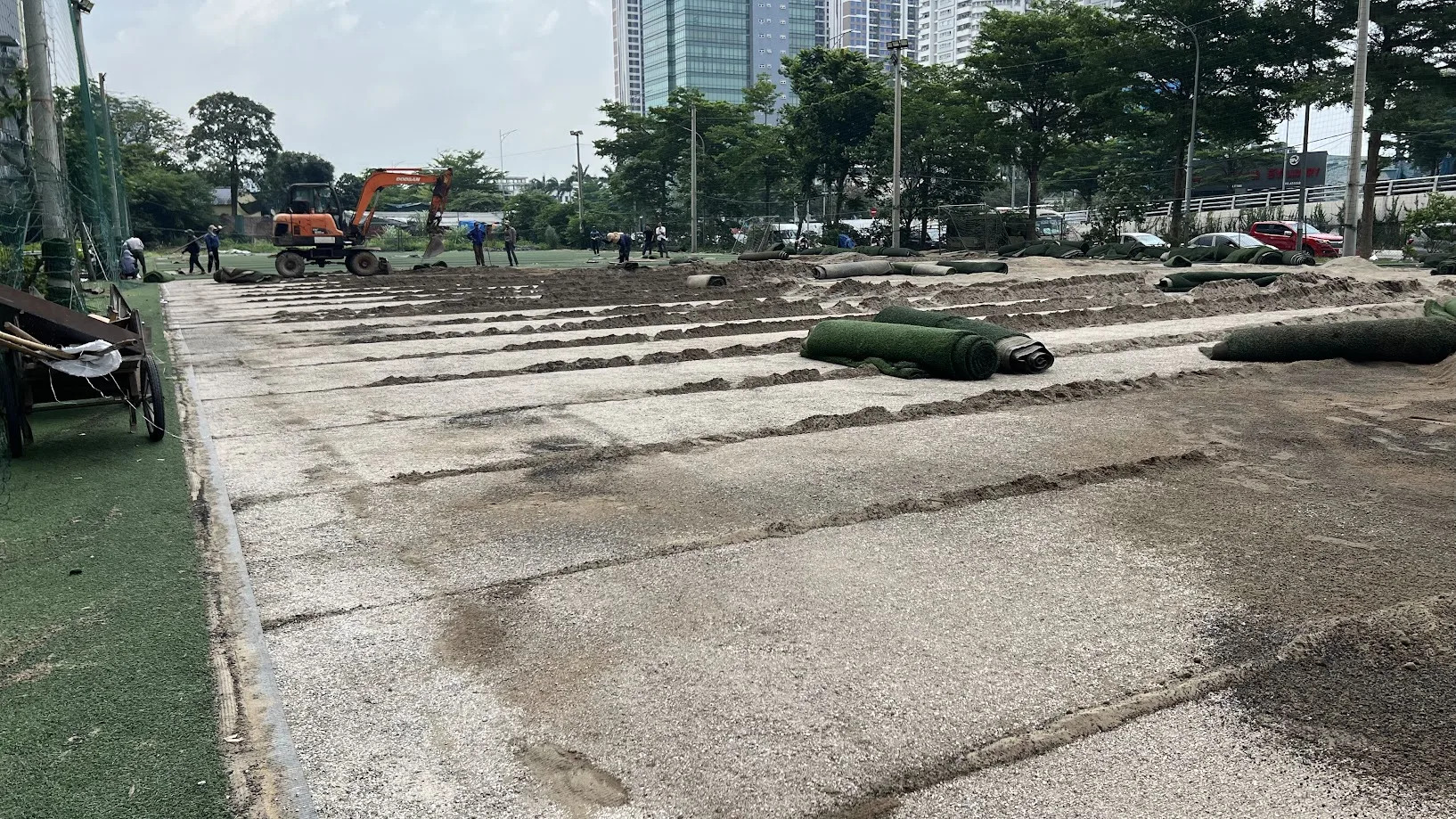 Đội thi công đang cuộn bóc lớp cỏ cũ khỏi mặt sân, cuộn cỏ được xếp gọn gàng